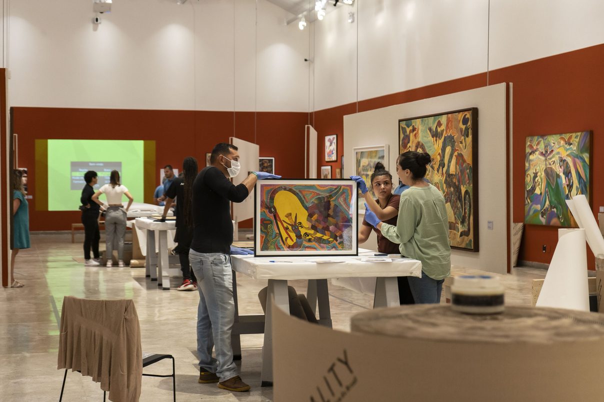 A team handles a framed painting on a table in a terracotta-walled gallery, other works hanging around them. Foto: Marília Camelo/Pinacoteca do Ceará