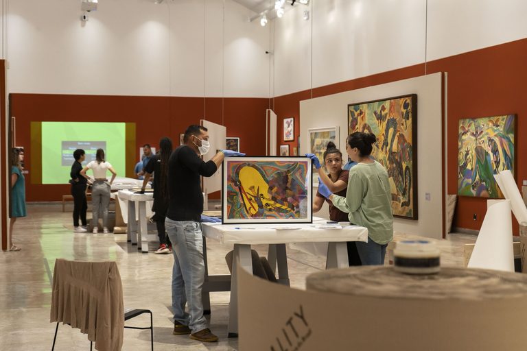 A team handles a framed painting on a table in a terracotta-walled gallery, other works hanging around them. Foto: Marília Camelo/Pinacoteca do Ceará
