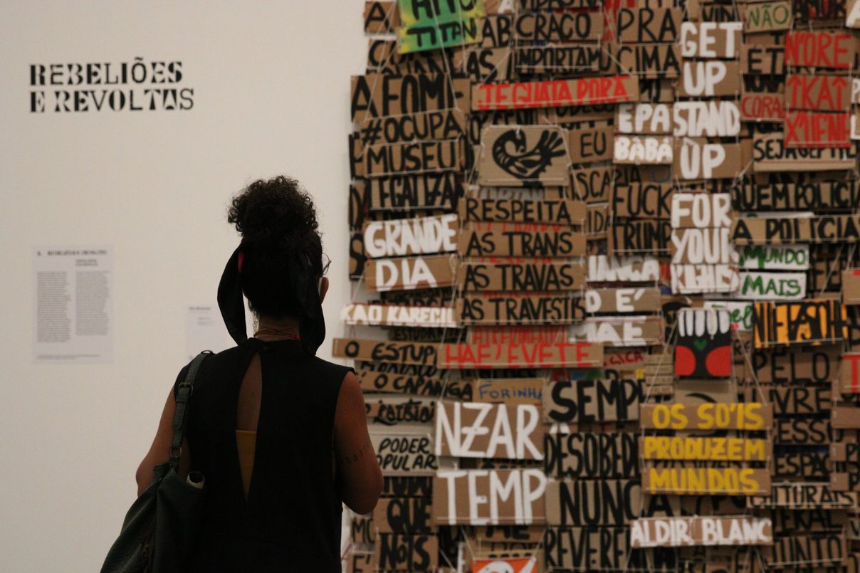 A visitor seen from behind facing a wall covered in hand-painted wooden signs, beside the panel Rebeliões e Revoltas. Foto: Rovena Rosa/Agência Brasil