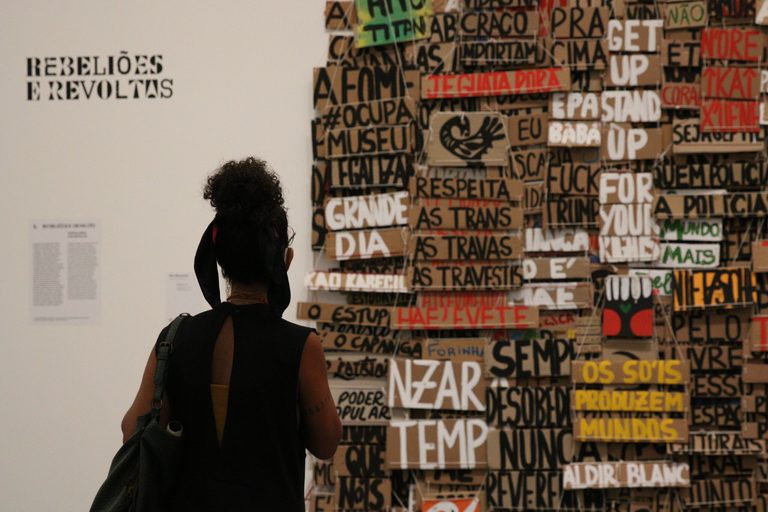 A visitor seen from behind facing a wall covered in hand-painted wooden signs, beside the panel Rebeliões e Revoltas. Foto: Rovena Rosa/Agência Brasil