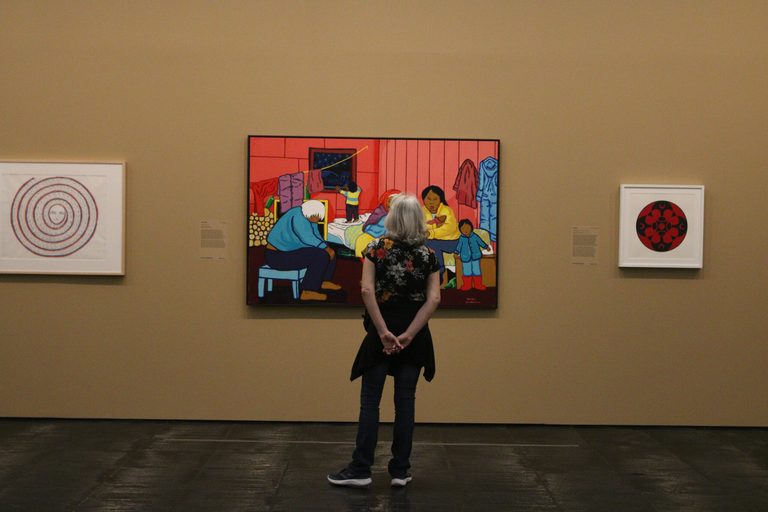 A visitor seen from behind before a large colourful painting on an ochre wall, flanked by two smaller works. Foto: Rovena Rosa/Agência Brasil