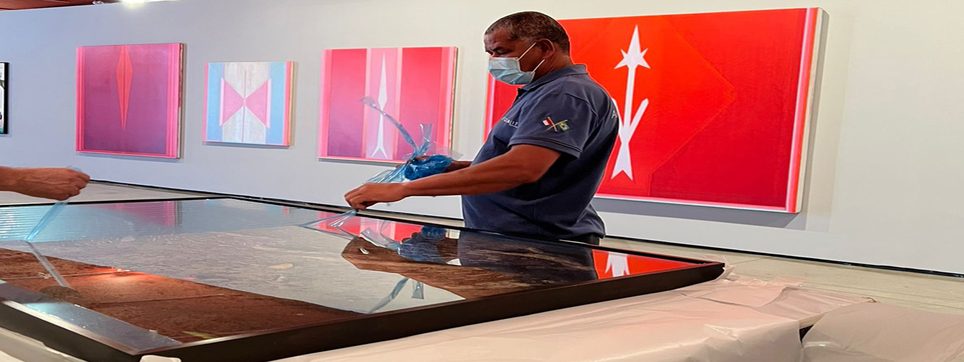 Funcionário de máscara e luvas manuseia uma placa de vidro sobre uma mesa, diante de pinturas vermelhas e rosas