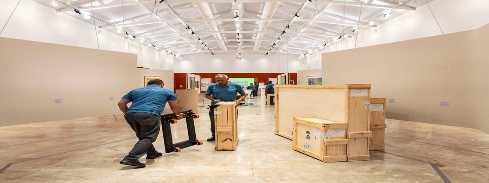 Dois funcionários da ArtQuality atravessam a galeria de um museu empurrando caixas de madeira em carrinhos