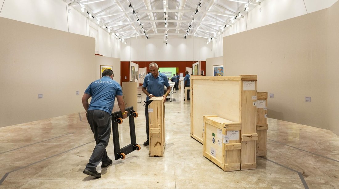 Dois funcionários da ArtQuality conduzem caixas de madeira em carrinhos por um corredor de exposição iluminado. Foto: Marília Camelo/Pinacoteca do Ceará
