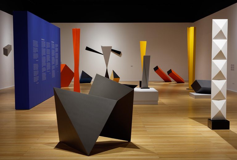 Exhibition room with geometric sculptures in blue, red, yellow and black on a wooden floor. Foto: Jaime Acioly