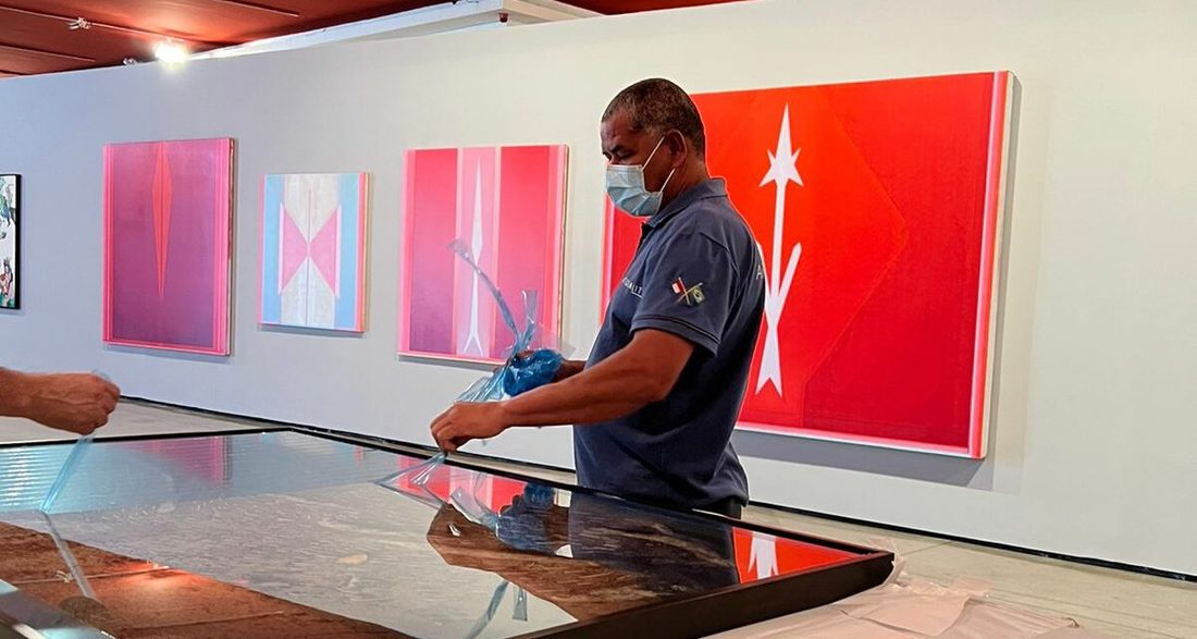 Funcionário de máscara e luvas apoia uma obra emoldurada sobre a mesa, numa sala com pinturas vermelhas ao fundo