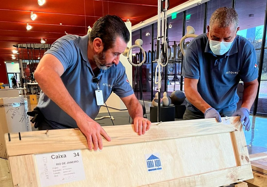 Deux manutentionnaires en chemise bleue ouvrent une caisse en bois étiquetée Caixa 34 dans une galerie éclairée en rouge
